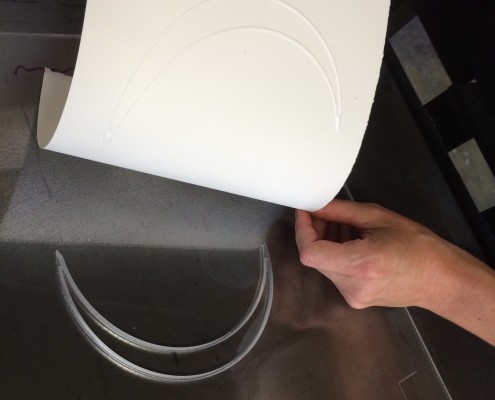 Crescent print and printing plate for Jaynes's The Moon Reader, 2014. Picture shows a translucent printing plate with the image of a crescent moon deeply etched into it. A white piece of paper with the crescent image embossed on it is being peeled away by the disembodied pair of hands of the printer. Her right hand, farthest from the viewer, holds the paper in the upper right corner. Her left hand, close to the viewer, is above the lower right edge of the bottom of the paper still attached to the plate. The reflection of the woman printer, who wears glasses, is seen in the lower edge of the printing plate, which is closest to the viewer. [End of description]