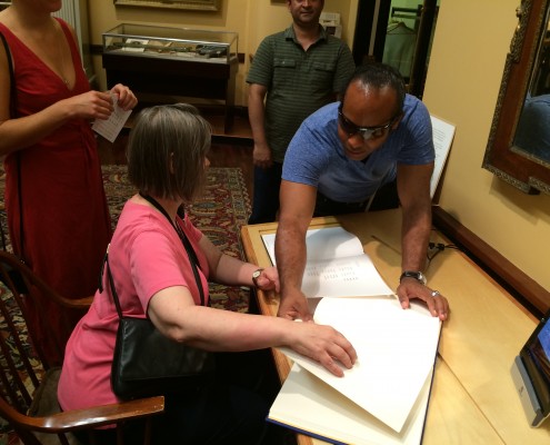 The Moon Reader advisors Suzanne Erb and Edery Herrera, Library Company opening, 2014. Picture shows Erb seated at a wooden-topped desk. An open bound volume of white pages, The Moon Reader, is on the right edge of the desk closest to the viewer. The Reader is below a tablet screen positioned upright in a stand. Another bound volume open to a page with printed text on white paper is to the left of The Moon Reader. Erb has her right hand on the page of the Reader and her left hand resting below the other bound volume. Herrera leans over the desk from Erb's left side and touches the Reader. A woman stands behind Erb, visible over her shoulder, and a man stands behind Hererra, visible over his shoulder. A display case of artifacts stands in the background. [End of description]