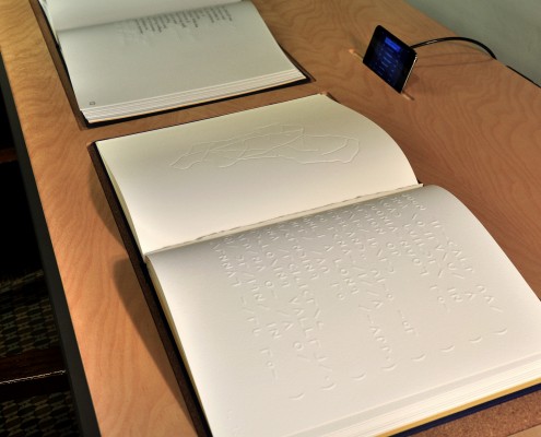 Teresa Jaynes, The Moon Reader, 2014. Picture depicts two large, open books displayed side by side on a wooden table. Above and behind the books, a black iPod is propped up in a small slot, and a black cord, connected to the iPod, extends down behind the table and out of view. The book closest to the viewer is a tactile book, opened to show, on the right page, a raised line map of a country or continent with divided sections. The left page shows a reverse imprint of Moon type symbols, embossed on the page before it. A page has been torn out of the book, leaving only a short, ragged edge of the page along the center fold. A purple or royal blue cloth binding with yellow endpaper is visible around the edge of the book’s pages. In the book farthest from the viewer, the right page displays a black non-tactile line drawing of the same map, while the left page is blank. Both books have white, clean paper and appear to be in excellent condition. [End of description]