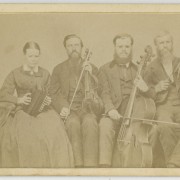 [Portrait of Hostetler family of blind musicians]. Mount Pleasant, Penna.: From A. N. Staufer, [ca. 1866]. Black & white photograph. 4 x 2.5 in. Picture shows one woman and three men, seated next to each other, and holding instruments. The woman holds an accordion in her lap and she looks slightly down. To her left is a man, his eyes closed, who holds a viola perpendicular to his lap with one hand and a bow in his other. To his left is a man resting a cello between his legs. He holds a bow across the base of the cello with his right hand. To his left is the last man, his eyes closed, who holds a violin by his left shoulder and a raised bow in in his right hand. The woman, as well as the man who holds a cello, wear glasses. The woman wears a dark-colored corseted dress with long sleeves and a long skirt. The men, who look toward the viewer, are bearded and wear dark-colored suits. [End of description]]