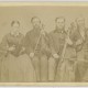 [Portrait of Hostetler family of blind musicians]. Mount Pleasant, Penna.: From A. N. Staufer, [ca. 1866]. Black & white photograph. 4 x 2.5 in. Picture shows one woman and three men, seated next to each other, and holding instruments. The woman holds an accordion in her lap and she looks slightly down. To her left is a man, his eyes closed, who holds a viola perpendicular to his lap with one hand and a bow in his other. To his left is a man resting a cello between his legs. He holds a bow across the base of the cello with his right hand. To his left is the last man, his eyes closed, who holds a violin by his left shoulder and a raised bow in in his right hand. The woman, as well as the man who holds a cello, wear glasses. The woman wears a dark-colored corseted dress with long sleeves and a long skirt. The men, who look toward the viewer, are bearded and wear dark-colored suits. [End of description]]