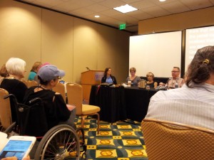 Picture shows view of a panel of people from the perspective of an audience member. Three women and a man sit at a table covered with a black fitted tablecloth. A paper coffee cup with lid, metal water pitcher, a pile of lanyards, and a water bottle are on the table in front of the panelists. Two screens, including one with closed captioned text, and a podium pushed against the wall are visible. In the foreground, audience members, including a female wheelchair user, are seated. The room contains beige paneled walls and patterned carpeting.