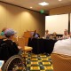 Picture shows view of a panel of people from the perspective of an audience member. Three women and a man sit at a table covered with a black fitted tablecloth. A paper coffee cup with lid, metal water pitcher, a pile of lanyards, and a water bottle are on the table in front of the panelists. Two screens, including one with closed captioned text, and a podium pushed against the wall are visible. In the foreground, audience members, including a female wheelchair user, are seated. The room contains beige paneled walls and patterned carpeting.