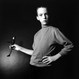 Picture shows a lanky older boy from his upper legs up. He stands, his arms posed, and looking directly at the viewer. His hair is combed severely back from his face. His body is slightly angled away from the viewer. The left part of his face is partly in shadow. His left arm is bent at the elbow and his left hand rests on his hip. His right arm lies close to his body. It is bent at the elbow at a right angle to his body. His right hand is extended and he holds a Catwoman doll in a similar pose to himself. He wears a dark-colored turtleneck, with the sleeves slightly pushed up, and black pants. [end of description]