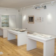 Picture shows exhibition installation shot of Touching the Book: Embossed LIterature for Blind People, taken from the back of the room and facing the exhibition entrance at the further end of the room on the right hand side. The flooring is light wood, and the walls are painted white. The photograph shows three free-standing display cases all containing 4-5 books opened and resting on cradles; two framed items on the right hand side of the wall as well as a large vinyl graphic of Moon's alphabetic system; and information panels are installed on the wall. At the further end of the room, and to the left of the entrance, is a large fixed display cabinet with three shelves holding artifacts (books and a writing frame). To the left of the cabinet is a blank TV screen and below that a table with a red covering that has been laid out with glasses of wine. [end of description]