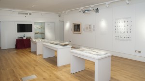 Picture shows exhibition installation shot of Touching the Book: Embossed LIterature for Blind People, taken from the back of the room and facing the exhibition entrance at the further end of the room on the right hand side. The flooring is light wood, and the walls are painted white. The photograph shows three free-standing display cases all containing 4-5 books opened and resting on cradles; two framed items on the right hand side of the wall as well as a large vinyl graphic of Moon's alphabetic system; and information panels are installed on the wall. At the further end of the room, and to the left of the entrance, is a large fixed display cabinet with three shelves holding artifacts (books and a writing frame). To the left of the cabinet is a blank TV screen and below that a table with a red covering that has been laid out with glasses of wine. [end of description]