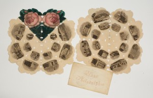 Picture shows two flower-shaped prints of “eight petals” illustrated with vignettes of buildings, landscape views, and portraits arranged in a circle of images nested within an outer circle of images. Very tiny rectangular and triangular shaped cut-outs line the open folds of the print. The jagged-edged prints are accompanied by an envelope printed in cursive letters in red ink reading: “Rose of Philadelphia.” The print in the left also contains two images of a pink rose bud with green petals as “petals” on its upper edge. [End of description]