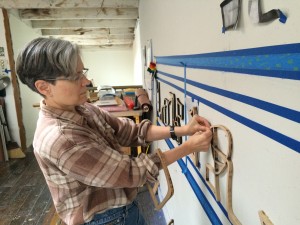 Picture shows Teresa Jaynes at work in her studio. Jaynes stands to the viewer's left. She looks at her hands as she adjusts a large-size, wooden sculptural letter between lines of blue tape on a white wall.