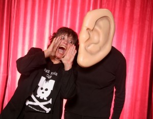 Picture shows Terry Galloway, standing and tilted to her left and with her hands up to her wide-open mouth.She shouts into a large -size ear that is the "face" of a person dressed all in black. A red curtain adorns the background.