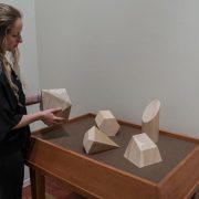Picture shows a woman, in the far left, holding one of five differently-shaped wooden geometric forms that rest on a wooden table. The top is covered in brown linen. Shapes include a trapezoid, trapezium, octagon, slant and flat-edged cylinder, and prism. The woman wears a black shirt and pants, and has her long blond hair pulled slightly away from her face.