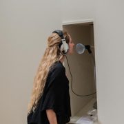 Picture shows a woman standing in front of a niche in a wall. She wears a headset, its cord extending from within the nice. Her face is near a funnel at the end of a mic-like stand. She wears a black shirt and pants, and has her long blond hair pulled slightly away from her face.