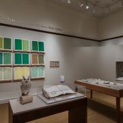 Picture shows the gallery, including four of Jaynes’s seven installations on view. In the center foreground is Jaynes’s Gift#1 on display on a flat, horizontally-angled rectangular table with a dark wood base. A white and brown paper owl rests on a cylindrical stand to the left of a large open book under a rectangular, clear acrylic hood. To the right of the display with the owl stands Jayne’s Gift #5, a map representation of the travels the 19th–century blind surveyor John Metcalf. It is a vertically-positioned, large rectangular table composed of a a linen top and a light brown wood base. A multi-color grid, outlines of geometric shapes, and green porcelain geometric shapes adorn the linen top. In the center background, on the back wall painted off-white, is Jayne’s Gift #4, a visual transmutation after the musical work of blind African American musician Thomas Wiggins. It is three horizontal and three vertical rows of prints in a geometric interplay of greens, browns and yellows. To the right of the prints on the wall is a small wooden frame in which brass musical notes are displayed. To the far right background is a view of Jaynes’s Gift #6, the scent mechanism the olfactometer, in a niche in an off-white curved wall. The floor of the room is covered with a tan colored carpet.