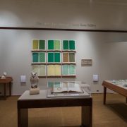 Picture shows the majority of the gallery, including four of Jaynes’s seven installations on view. In the center foreground is Jaynes’s Gift#1 on display on a flat, horizantally-angled rectangular table with a dark wood base. A white and brown paper maiche owl rests on a cylindrical stand to the left of a large open book under a rectangular, clear acrylic hood. To the right of the display with the owl stands Jayne’s Gift #5, a map represention of the travels of the 19th–century blind surveyor John Metcalf. It is a vertically-positioned, large rectangular table composed of a a padded linen top and a light brown wood base. A multi-color grid, outlines of geometric shapes, and green porcelain geometric forms adorn the linen top. To the left of the owl display is a partial view of a case of historical materials. In the left backround is Jayne’s Gift #3 inspired by the mathematical tools of the blind mathematician Nicholas Saunderson. It is a light brown table on which several large-sized wooden geometric shapes of different styles rest. In the center background, on the back wall painted off-white, is Jayne’s Gift #4, a visual transmutation after the musical work of blind African Amerian musician Thomas Wiggins. It is three horizontal and three vertical rows of prints in a geometric interplay of greens, browns and yellows. To the right of the prints on the wall is a small wooden frame in which brass musical notes are displayed. To the far right background is a partial view of an off-white curved wall. The floor of the room is covered with a tan colored carpet.