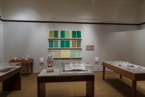 Picture shows the majority of the gallery, including four of Jaynes’s seven installations on view. In the center foreground is Jaynes’s Gift#1 on display on a flat, horizantally-angled rectangular table with a dark wood base. A white and brown paper maiche owl rests on a cylindrical stand to the left of a large open book under a rectangular, clear acrylic hood. To the right of the display with the owl stands Jayne’s Gift #5, a map represention of the travels of the 19th–century blind surveyor John Metcalf. It is a vertically-positioned, large rectangular table composed of a a padded linen top and a light brown wood base. A multi-color grid, outlines of geometric shapes, and green porcelain geometric forms adorn the linen top. To the left of the owl display is a partial view of a case of historical materials. In the left backround is Jayne’s Gift #3 inspired by the mathematical tools of the blind mathematician Nicholas Saunderson. It is a light brown table on which several large-sized wooden geometric shapes of different styles rest. In the center background, on the back wall painted off-white, is Jayne’s Gift #4, a visual transmutation after the musical work of blind African Amerian musician Thomas Wiggins. It is three horizontal and three vertical rows of prints in a geometric interplay of greens, browns and yellows. To the right of the prints on the wall is a small wooden frame in which brass musical notes are displayed. To the far right background is a partial view of an off-white curved wall. The floor of the room is covered with a tan colored carpet.