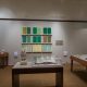 Picture shows the majority of the gallery, including four of Jaynes’s seven installations on view. In the center foreground is Jaynes’s Gift#1 on display on a flat, horizantally-angled rectangular table with a dark wood base. A white and brown paper maiche owl rests on a cylindrical stand to the left of a large open book under a rectangular, clear acrylic hood. To the right of the display with the owl stands Jayne’s Gift #5, a map represention of the travels of the 19th–century blind surveyor John Metcalf. It is a vertically-positioned, large rectangular table composed of a a padded linen top and a light brown wood base. A multi-color grid, outlines of geometric shapes, and green porcelain geometric forms adorn the linen top. To the left of the owl display is a partial view of a case of historical materials. In the left backround is Jayne’s Gift #3 inspired by the mathematical tools of the blind mathematician Nicholas Saunderson. It is a light brown table on which several large-sized wooden geometric shapes of different styles rest. In the center background, on the back wall painted off-white, is Jayne’s Gift #4, a visual transmutation after the musical work of blind African Amerian musician Thomas Wiggins. It is three horizontal and three vertical rows of prints in a geometric interplay of greens, browns and yellows. To the right of the prints on the wall is a small wooden frame in which brass musical notes are displayed. To the far right background is a partial view of an off-white curved wall. The floor of the room is covered with a tan colored carpet.