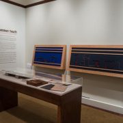 Picture shows Jaynes’s Gift #2, an interactive piece inspired by the physicality of the wooden writing frames and the abstract quality of letterforms written in the 1870s by Jennie Partridge who was blind. It is two oversized, horizontal writing frames, with felt backgrounds, mounted alongside one another on an off-white painted wall. Beneath the top of the frame are two evenly spaced horizontal wooden strips. Then there is a wider space and another horizontal strip. The left frame, with blue felt, serves as a mount for free standing, abstract letterforms constructed in darkly painted wood. The letters in the large frame on the right, with black felt, are more formally set within their indented image. The elaborate, enlarged letter forms spell out the phrase: “I know.” In the foreground is a case with historical materials. In the left background is the introductory text to the exhibition.