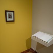 Picture shows a yellow painted wall on which only a wooden frame with a late 19th-century black and white cabinet card portrait hangs in the center. The portrait shows a couple. To the lower right is an open book of white pages, Jayne’s Gift #7, resting on a pedestal that juts from an off-white painted wall.