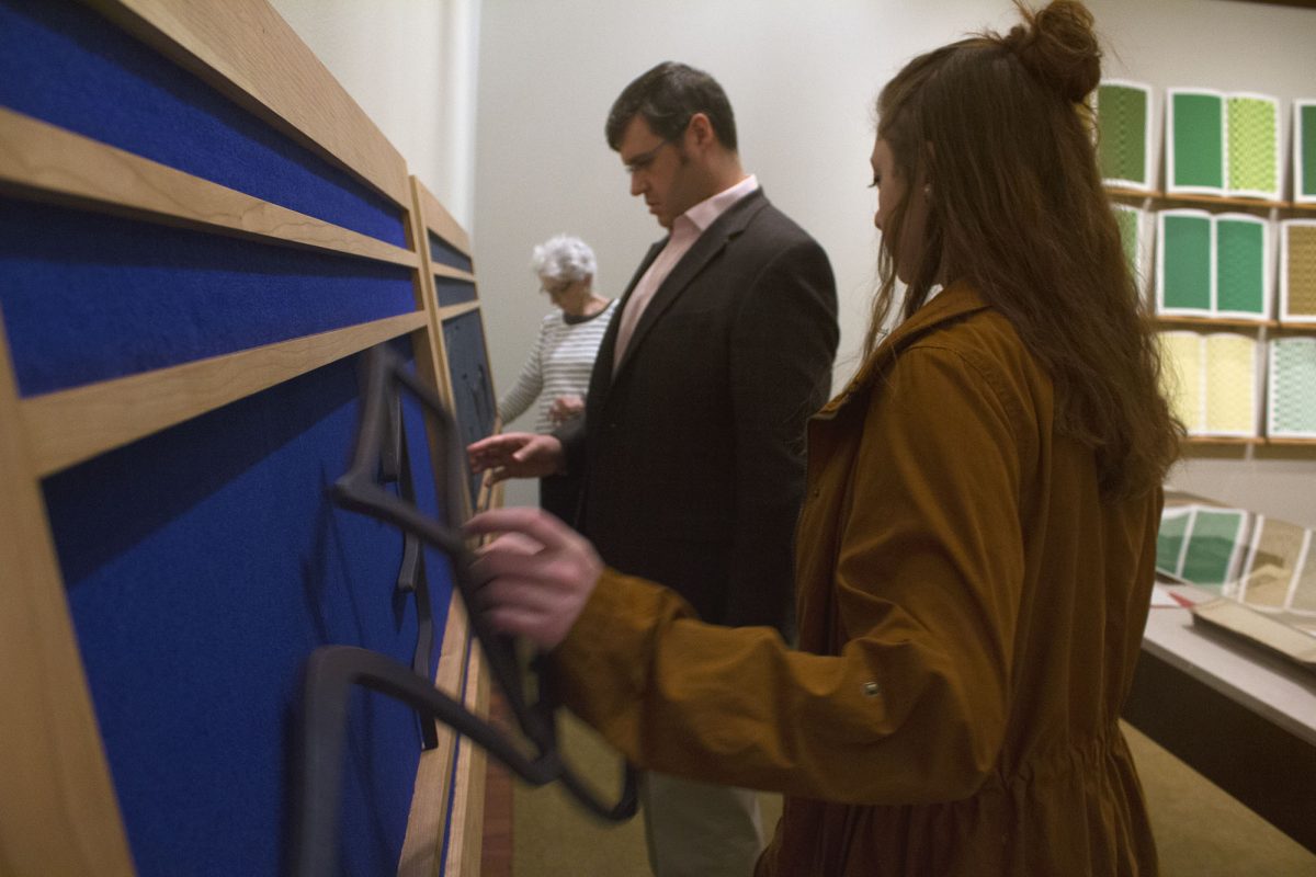 Picture shows a man and woman touching the sculptural letters and two large frames of Gift #2. A woman, with long dark hair and a tan coat, holds a large sculptural letter from the frame closer to the viewer in her left hand. To her right, a man in a grey suit jacket touches the felt covering of the frame farthest from the viewer. In the background, the screen prints of Jaynes’s Gift #4 and another woman visitor is visible. [end of description]