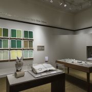Picture shows the gallery, including four of Jaynes’s seven installations on view. In the center foreground is Jaynes’s Gift#1 on display on a flat, horizontally-angled rectangular table with a dark wood base. A white and brown paper owl rests on a cylindrical stand to the left of a large open book under a rectangular, clear acrylic hood. To the right of the display with the owl stands Jayne’s Gift #5, a map representation of the travels the 19th–century blind surveyor John Metcalf. It is a vertically-positioned, large rectangular table composed of a a linen top and a light brown wood base. A multi-color grid, outlines of geometric shapes, and green porcelain geometric shapes adorn the linen top. In the center background, on the back wall painted off-white, is Jayne’s Gift #4, a visual transmutation after the musical work of blind African American musician Thomas Wiggins. It is three horizontal and three vertical rows of prints in a geometric interplay of greens, browns and yellows. To the right of the prints on the wall is a small wooden frame in which brass musical notes are displayed. To the far right background is a view of Jaynes’s Gift #6, the scent mechanism the olfactometer, in a niche in an off-white curved wall. The floor of the room is covered with a tan colored carpet. [end of description]