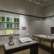 Picture shows the gallery, including four of Jaynes’s seven installations on view. In the center foreground is Jaynes’s Gift#1 on display on a flat, horizontally-angled rectangular table with a dark wood base. A white and brown paper owl rests on a cylindrical stand to the left of a large open book under a rectangular, clear acrylic hood. To the right of the display with the owl stands Jayne’s Gift #5, a map representation of the travels the 19th–century blind surveyor John Metcalf. It is a vertically-positioned, large rectangular table composed of a a linen top and a light brown wood base. A multi-color grid, outlines of geometric shapes, and green porcelain geometric shapes adorn the linen top. In the center background, on the back wall painted off-white, is Jayne’s Gift #4, a visual transmutation after the musical work of blind African American musician Thomas Wiggins. It is three horizontal and three vertical rows of prints in a geometric interplay of greens, browns and yellows. To the right of the prints on the wall is a small wooden frame in which brass musical notes are displayed. To the far right background is a view of Jaynes’s Gift #6, the scent mechanism the olfactometer, in a niche in an off-white curved wall. The floor of the room is covered with a tan colored carpet. [end of description]