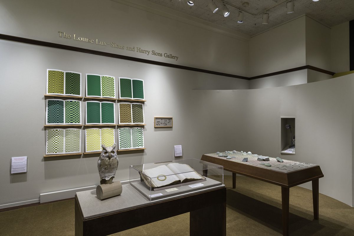 Picture shows the gallery, including four of Jaynes’s seven installations on view. In the center foreground is Jaynes’s Gift#1 on display on a flat, horizontally-angled rectangular table with a dark wood base. A white and brown paper owl rests on a cylindrical stand to the left of a large open book under a rectangular, clear acrylic hood. To the right of the display with the owl stands Jayne’s Gift #5, a map representation of the travels the 19th–century blind surveyor John Metcalf. It is a vertically-positioned, large rectangular table composed of a a linen top and a light brown wood base. A multi-color grid, outlines of geometric shapes, and green porcelain geometric shapes adorn the linen top. In the center background, on the back wall painted off-white, is Jayne’s Gift #4, a visual transmutation after the musical work of blind African American musician Thomas Wiggins. It is three horizontal and three vertical rows of prints in a geometric interplay of greens, browns and yellows. To the right of the prints on the wall is a small wooden frame in which brass musical notes are displayed. To the far right background is a view of Jaynes’s Gift #6, the scent mechanism the olfactometer, in a niche in an off-white curved wall. The floor of the room is covered with a tan colored carpet. [end of description]