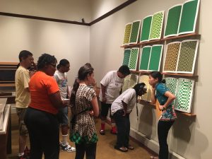 Photo shows a large group of individuals gathered in front of Teresa Jaynes’s Gift #4 of nine green, yellow, and brown screen prints. The screen prints depict grid patterns. In the right of the image, a young woman attired in black pants and a short-sleeved teal blouse with a flower pattern on the edge, slightly hunches over as if listening to something. Her right hand is to her ear and her left hand holds a large envelope to her waist. To her left, are two women. They lean and bend over in listening stances. In the left of the image, is a group of men and women in summer clothes. They look toward the women standing in front of the screen prints. The screen prints are hung above a short white-colored floor riser. [end of description]